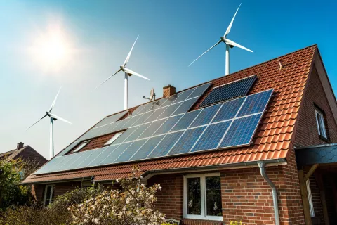 Ein Hausdach, das Sonnenkollektoren installiert hat. Im Hintergrund sind Windkrafträder und blauer Himmel.